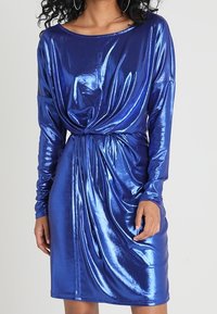 Lesklé modré šaty s dlouhými rukávy, plisovaným předním dílem a zkrouceným uzlem v pase. Hladká, lesklá látka s přiléhavým střihem.