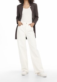 Pantalones amplios blancos combinados con una camiseta sin mangas blanca ajustada, debajo de una chaqueta larga marrón oscuro. La modelo está de pie con las manos en los bolsillos.