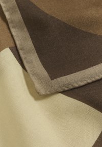 Tissu en coton avec des motifs géométriques en brun, beige et jaune. Coutures remarquables le long des bords. Surface texturée visible.