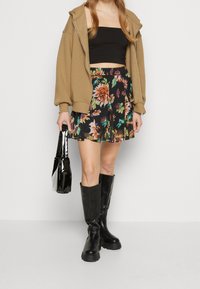 Crop top noir, sweat à capuche beige surdimensionné, jupe florale avec des verts et des roses, bottes noires jusqu'aux genoux, et un sac à main noir brillant.
