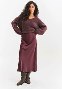 Žena s dlouhými kudrnatými vlasy, oblečená v nadýchaném fialovém svetru, ladící midi sukni z lesklého saténu a hnědých botách sahajících do poloviny lýtek, stojící s rukama v bok.