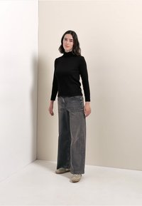 Maglietta a dolcevita nera aderente abbinata a jeans in denim grigio a gamba larga. La modella si trova su uno sfondo neutro, indossando scarpe di colore chiaro.