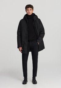 Giacca a vento nera con cappuccio, chiusura con zip e tasche laterali, indossata sopra un dolcevita nero e abbinata a pantaloni neri slim e scarpe.