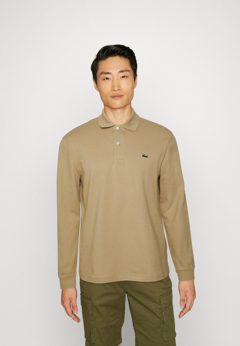 Beiges Langarm-Poloshirt mit weicher Textur, zwei Knöpfen am Kragen und einem grünen Lacoste-Logo auf der linken Brust. Lässige Passform.