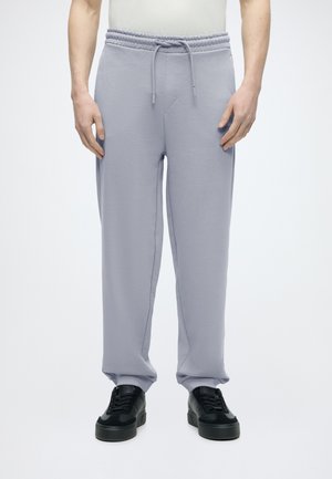 Mand iført lysegrå joggingbukser med snøre i taljen og sorte sneakers, stående mod en ensfarvet lys baggrund.