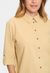 Light yellow, cotton button-up shirt with a collar, three-quarter sleeves, and brown buttons. Soft texture with rolled sleeve cuffs.