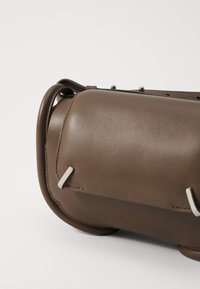Geantă din piele maro, cu o formă rotunjită, având accesorii din metal argintiu și cusături detaliate. Textură netedă cu accente proeminente.