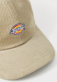 Beige kordfløyelslue med ribbet tekstur, med en brodert Dickies-logo i rødt, gult og blått på fronten. Skjermen er lett buet.
