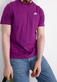 Lila bomull T-shirt med rund halsringning och korta ärmar. Har en vit Nike-logotyp på bröstet. Hålls med en hand, ihop med blå jeans.