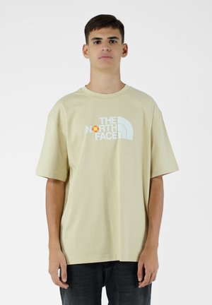 Camiseta de algodón beige con un logo blanco y acentos en naranja y amarillo. Manga corta, corte holgado, diseño de cuello redondo. Textura simple y suave.