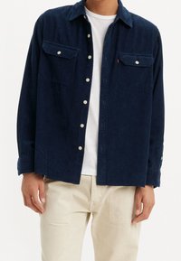 Marinblå manchester skjorta med knäppta bröstfickor, vita knappar och krage. Bärs över en vit T-shirt, kombinerad med ljusfärgade byxor.