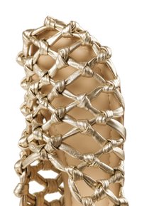 Κοντινό πλάνο στο δάχτυλο ενός παπουτσιού με μεταλλικούς χρυσούς δερμάτινους ιμάντες που διασταυρώνονται και δένονται σε κόμπους σχηματίζοντας ένα ανοιχτό πλέγμα.