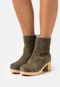 Botas de tobillo de ante verde oliva con un cuello de piel sintética, tacón de bloque de madera y costuras decorativas a lo largo de los bordes.