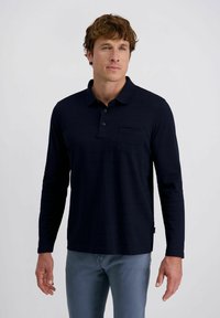 Mann mit lockigem braunem Haar, der ein schwarzes Langarm-Polohemd und graue Hosen trägt, vor einem einfarbigen hellen Hintergrund stehend.