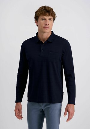 Mann mit lockigem braunem Haar, der ein schwarzes Langarm-Polohemd und graue Hosen trägt, vor einem einfarbigen hellen Hintergrund stehend.