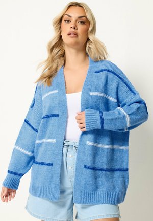 Lyshåret kvinde iført oversized blå strikket cardigan med hvide og marineblå striber, hvid top og lyse denimshorts mod ensfarvet baggrund.