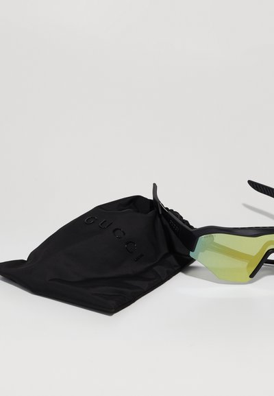 Óculos de sol pretos com um design angular, lentes com tonalidade verde, presos a um saco de tecido preto rotulado "GUCCI."