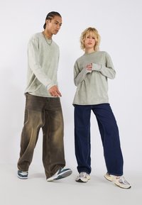 To personer har på seg dempede langermede skjorter i grønn og grå, kombinert med løse jeans - en brun falmet og en mørk blå. Begge har sneakers på føttene.
