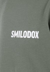 Camiseta de algodón verde oliva con el texto blanco "SMILODOX" exhibido de manera prominente. Textura suave, mangas cortas y corte casual.