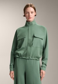 Giacca cropped verde con colletto alto realizzata in tessuto morbido, dotata di due tasche frontali, polsini a coste e orlo elastico per una vestibilità rilassata.