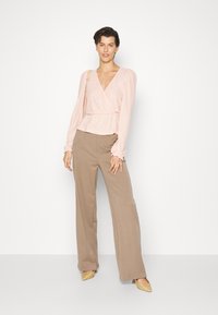 Blusa envolvente rosa claro com tecido texturizado e mangas longas, combinada com calças largas bege e sapatos amarelos de bico pontiagudo em palha.