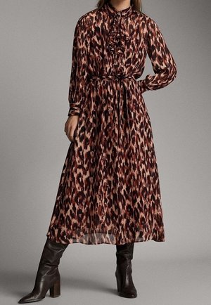 Femme portant une robe imprimée animal à manches longues et longueur cheville avec col à volants et bottes noires à talons, debout avec une main sur la hanche.