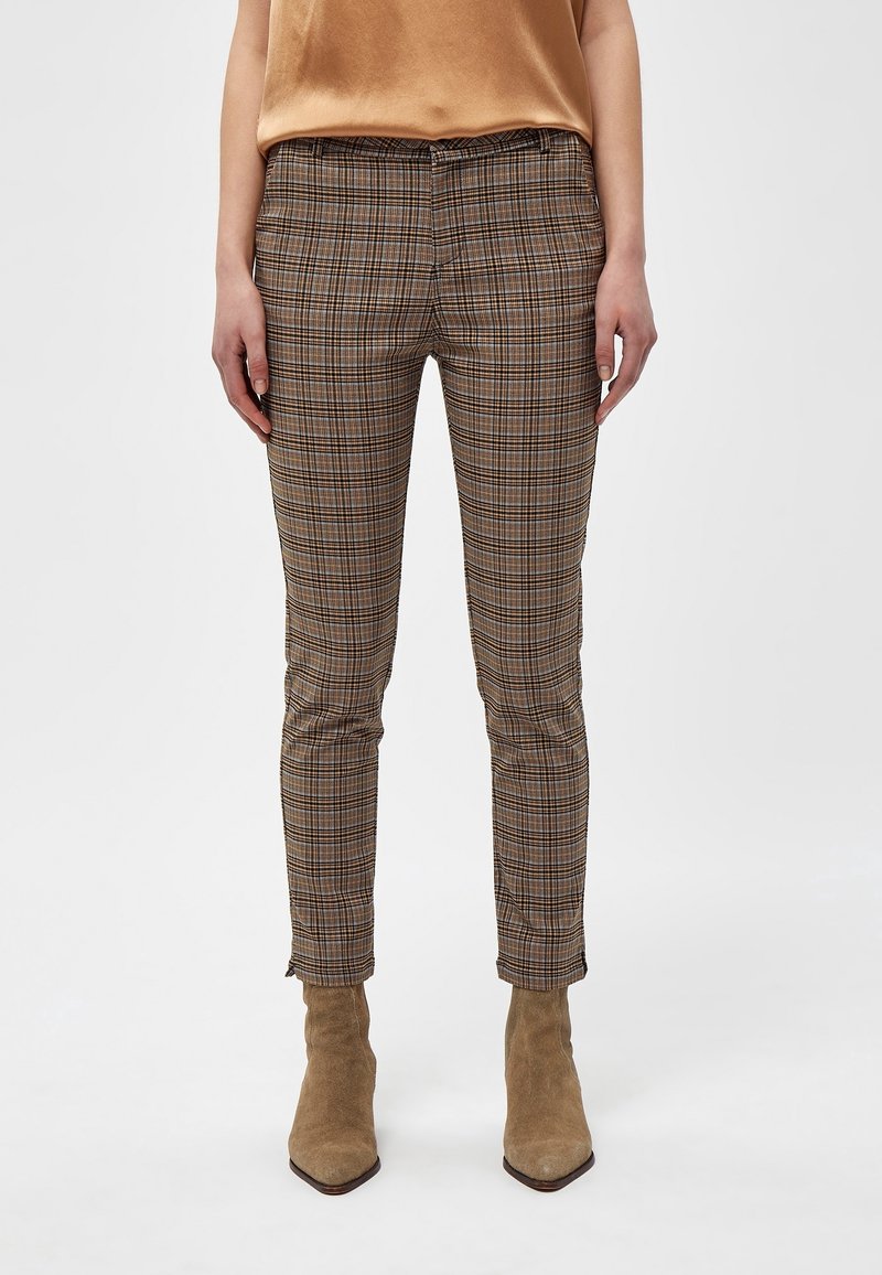 Femme portant un pantalon slim à carreaux marron, des bottines en daim couleur tan et un chemisier en satin couleur camel, debout devant un fond uni.