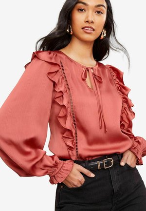 Rust-colored blouse with ruffled sleeves and a keyhole neckline, featuring vertical detailing and a textured fabric. Black high-waisted jeans.