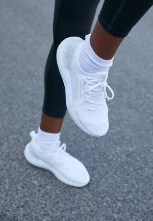 Jambes en leggings noirs avec des chaussettes blanches et des chaussures de sport blanches sur un sol gris texturé, un pied levé au milieu d'une foulée.