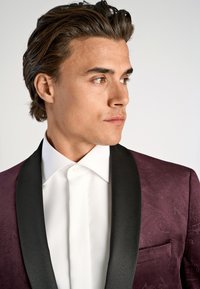 Burgundyröd blazer med strukturerat mönster, svart satinrevär och vit skjorta med en subtil strukturerad yta. Smal ficka på vänster sida.