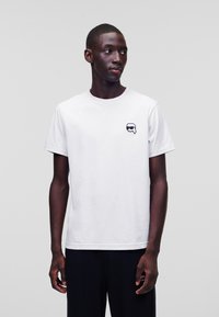 Balta medvilninė marškinė su trumpomis rankovėmis, apvaliu iškirpimu, su maža juoda grafika kairėje krūtinės pusėje. Paprastas, klasikinis dizainas.