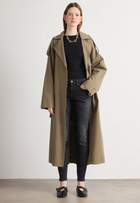 Hosszú olajzöld trench kabát fekete felső fölött, sötét skinny farmerrel és fekete lapos cipővel párosítva. Ezüst lánc nyaklánc és laza fit jellemzi.