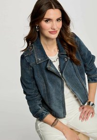 Giacca da motociclista in denim blu scuro con colletto strutturato, chiusura con zip e dettagli in metallo argentato. Il modello indossa una camicia bianca e pantaloni.