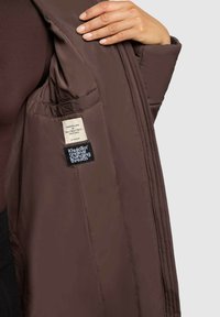 Jacheta izolată maro are un fermoar frontal, un interior quilted și două etichete în interior cu text. Exterior neted cu o strălucire moderată.