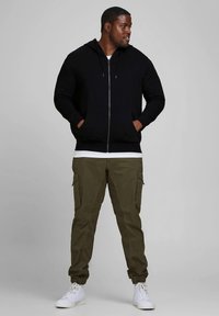 Melns rāvējslēdzēja džemperis ar priekšējām kabatām, kombinēts ar olīvu krāsas kravas biksēm ar sānu kabatām un sašaurinātām aprocēm, valkāts ar baltām sporta čībām.