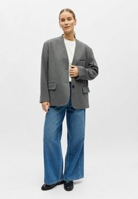 Blazer gris en mélange de laine avec un revers cranté, trois poches avant et une fermeture à deux boutons, associé à un jean bleu ample en denim et des chaussures noires.
