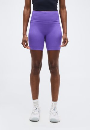 Γυναίκα που φοράει ποδηλατικά σορτς σε μωβ χρώμα με ριμπ ύφανση και ψηλή μέση, λευκές κάλτσες, λευκά αθλητικά παπούτσια και μαύρο κομψό μπουστάκι, στέκεται μπροστά σε απλό φόντο.