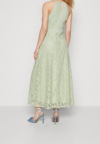 Ljusgrön spetsmaxiklänning med halterneck, som har en figurnära överdel och en flödande kjol. Matchad med blå högklackade skor.