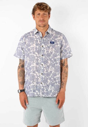 Chemise à manches courtes blanche avec un motif floral bleu, dotée de boutons et d'une poche poitrine avec une étiquette bleue. Portée avec un short clair.