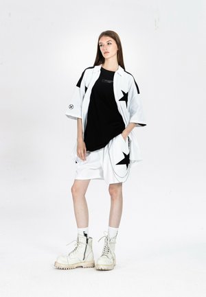 Mujer joven de pie vestida con pantalones cortos blancos de gran tamaño y camisa con estampados de estrellas negras, camiseta negra, calcetines blancos y botas blancas gruesas.