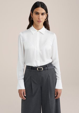Blusa blanca de manga larga con cuello abotonado, combinada con pantalones grises holgados y un cinturón de cuero negro con hebilla dorada.