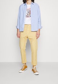 Chemise bleu clair à boutons, t-shirt blanc avec motif, pantalon jaune et baskets montantes marron. Look décontracté avec manches de chemise retroussées.