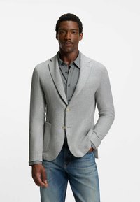 Grijze blazer van een gestructureerde stof, met een enkele knoopsluiting, twee voorzakken, gedragen over een patroonshirt, gecombineerd met blauwe jeans.