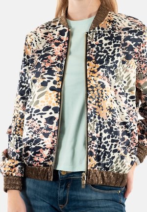 Vrouw die een multicolor bomberjack met vlekken en metalen bruine biezen draagt, over een lichtgroen shirt en blauwe spijkerbroek.