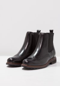 Bottes noires en cuir à la cheville avec détails brogue, panneaux latéraux élastiques et languettes de traction. Elles ont une semelle plate et un talon marron contrastant.