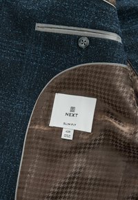 Inuti den blå tweedblazern finns en brun foder med ett subtilt hundtandsmönster. Taggen visar "NEXT", "SLIM FIT", storlek 40R.