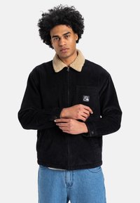 Sort fløjljakke med en beige sherpa-krave, lynlåslukning og en frontlomme med et logo. Modellen står med armene over kors.