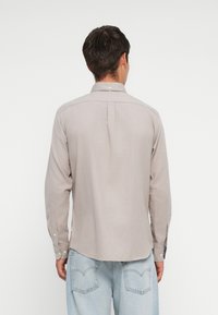 Svetlo šedá košeľa s dlhým rukávom, golierom, zapínaním na gombíky a zakriveným spodným okrajom. Obsahuje zúženie na chrbte pre väčšiu voľnosť pohybu.