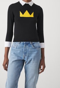 Pull en maille noire avec un motif de couronne jaune sur le devant, comprenant une chemise blanche à col en dessous et des manches trois-quarts.