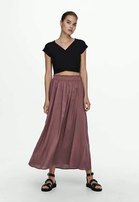 Jonge vrouw die staat, gekleed in een zwart kortemouw V-hals crop top, mauve high-waisted maxi rok en zwarte sandalen op een witte achtergrond.